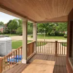 Brick house porch with a wood-plank ceiling, tan columns, and wooden railings.